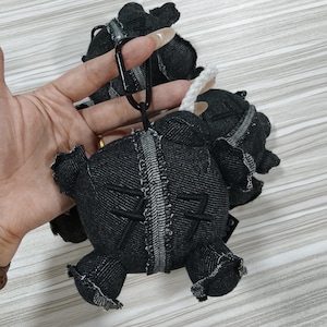 May include: Black denim elephant-shaped keychain with white stitching and a black carabiner clip. The elephant has small, ruffled ears and a textured denim appearance. The keychain is designed with a distressed aesthetic.