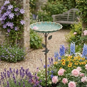 Puede incluir: Un ba&ntilde;o para p&aacute;jaros con un cuenco festoneado de color verde azulado y un interior texturizado con motivos florales. El cuenco se apoya en un soporte de metal marr&oacute;n oscuro adornado con hojas met&aacute;licas decorativas y una peque&ntilde;a figura de colibr&iacute;.