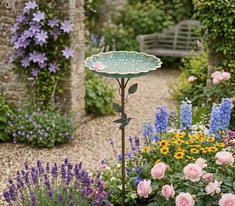 Puede incluir: Un ba&ntilde;o para p&aacute;jaros con un cuenco festoneado de color verde azulado y un interior texturizado con motivos florales. El cuenco se apoya en un soporte de metal marr&oacute;n oscuro adornado con hojas met&aacute;licas decorativas y una peque&ntilde;a figura de colibr&iacute;.