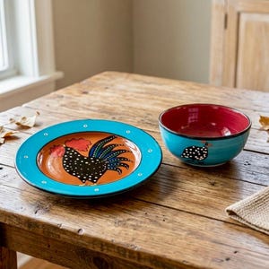 May include: A ceramic plate and bowl set featuring a hand-painted black rooster with white polka dots. The plate has a bright turquoise rim and an orange centre, while the bowl has a turquoise exterior and a deep red interior.