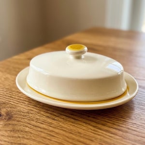May include: An oval ceramic butter dish featuring a smooth off-white lid with a small, raised mustard-yellow knob handle. The base plate is off-white with a distinct mustard-yellow band around the rim, matching the color of the lid handle.