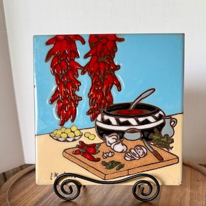 May include: A square ceramic tile featuring a painted scene of two bundles of red chilli peppers hanging against a light blue background. Below, a black and white patterned pot sits on a tan surface with garlic, sliced onions, and peppers.