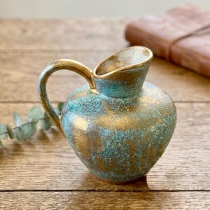 Może przedstawiać: Mały ceramiczny dzbanek z zaokrąglonym korpusem i zakrzywionym uchwytem. Powierzchnia ma teksturowane wykończenie z nakrapianą mieszanką metalicznego złota i turkusowego błękitu, tworząc efekt postarzenia z szerokim dzióbkiem do nalewania.