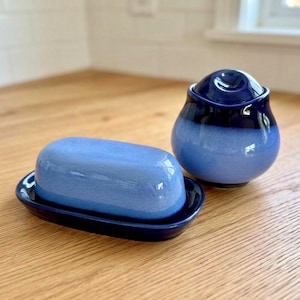 Könnte beinhalten: Eine Keramik-Butterdose mit einem hellblauen, gesprenkelten, ovalen Deckel und einem dunkelblauen Unterteil, begleitet von einer passenden dunkelblauen Zuckerdose mit hellblauem, gesprenkeltem, rundem Körper und einem dunkelblauen Deckel.