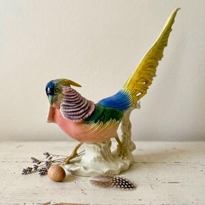 May include: A ceramic pheasant figurine featuring a vibrant color palette of pink, purple, blue, green, and yellow. The bird stands on a textured white base with a long, sweeping tail. A small wooden egg and a sprig of dried lavender are nearby.