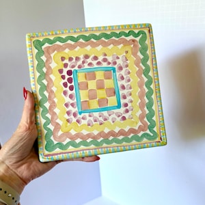 May include: A square ceramic tile featuring a hand-painted design with concentric borders of green, brown, and yellow wavy lines. The center displays a light brown and cream checkerboard pattern framed by a bright blue square and purple dots.