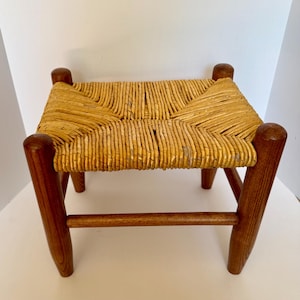 May include: A small rectangular stool featuring four cylindrical wooden legs connected by horizontal support bars. The seat is constructed from woven light brown natural fiber, creating a textured surface with a distinct diagonal pattern.