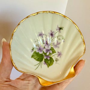 May include: A shell-shaped porcelain dish with a cream-colored surface featuring a central floral illustration of purple violets with green leaves. The scalloped edges of the dish are finished with a decorative gold trim along the entire rim.
