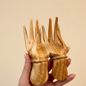 Può includere: Un pettine per capelli in legno marrone chiaro caratterizzato da un design originale di due mani intagliate con dita allungate che fungono da denti del pettine. La venatura naturale del legno è visibile sulla superficie liscia e lucida.
