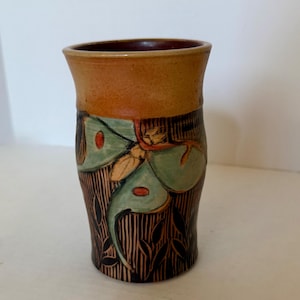 Op de afbeelding: Een cilindervormige keramische beker met een gladde, beige rand en een donkerbruine basis met verticale gegraveerde lijnen. Het lichaam toont een handgeschilderde lichtgroene nachtvlinder met oranje accenten en donkere bladvormige silhouetten.