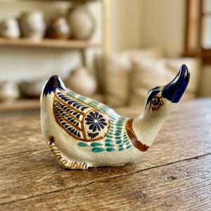 May include: A ceramic duck figurine featuring a cream-colored base with hand-painted details in dark blue, teal, and brown. The design includes intricate floral patterns on the wings, a solid blue beak, and a brown band around the neck.