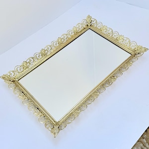 Puede incluir: Una bandeja rectangular con una superficie central de espejo encerrada por un marco de metal dorado ornamentado. El borde muestra intrincados patrones de volutas y filigrana con detalles en forma de hoja en cada esquina.