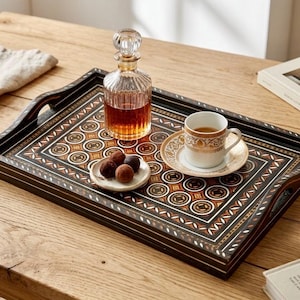 May include: A rectangular wooden serving tray featuring an intricate geometric inlay pattern in black, white, and brown. On the tray sits a clear glass decanter filled with amber liquid, a white ceramic cup and saucer, and a small plate with three truffles.