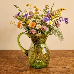 Peut inclure: Une carafe en verre vert transparent remplie d'un bouquet vibrant de fleurs sauvages, comprenant des boutons d'or jaunes, des bleuets, des marguerites blanches, du trèfle violet et des campanules, aux tiges vertes visibles à travers le verre.