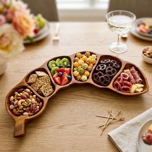 Könnte beinhalten: Ein geschwungenes, mehrteiliges Serviertablett aus Holz mit fünf separaten Fächern, gefüllt mit verschiedenen Snacks wie gemischten Nüssen, quadratischen Crackern, geschnittenen Kiwis und Erdbeeren, Blaubeeren, Popcorn, Schokoladenbrezeln und Aufschnitt.