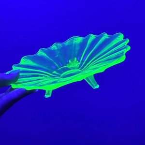 May include: A translucent, bright neon green glass bowl shaped like a scalloped shell. The bowl features a ribbed, fan-like design radiating from the center and is supported by three small, integrated glass feet, glowing intensely under UV light.