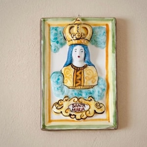 May include: A rectangular ceramic wall plaque featuring a relief of a crowned figure with blue hair and a yellow patterned garment. The figure is surrounded by light blue clouds, and a scroll at the bottom contains the text SANTA MARIA.