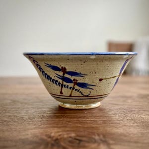 May include: A speckled ceramic bowl with a flared rim, featuring hand-painted blue dragonfly motifs and a thin dark brown line encircling the base. The exterior surface has a light beige, textured finish with subtle dark brown speckles throughout.