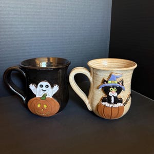 May include: Two ceramic mugs featuring Halloween designs. The left mug is black with a white ghost emerging from an orange pumpkin. The right mug is beige with a black cat wearing a purple witch hat, also sitting inside an orange pumpkin.