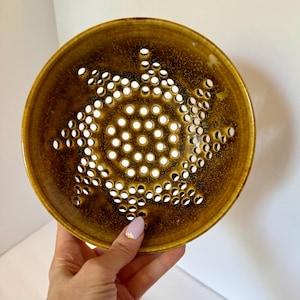 May include: A round, mustard-yellow ceramic bowl featuring a perforated design with numerous small, circular holes arranged in a spiral pattern radiating from the center. The glaze has a speckled, glossy finish with darker brown tones.