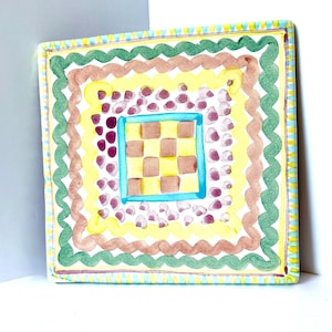 Può includere: Una piastrella quadrata in ceramica con un motivo a scacchiera centrale in giallo e marrone, racchiuso da un bordo blu brillante. Attorno si trovano strati concentrici di punti viola, una linea ondulata gialla, una linea ondulata marrone e un bordo verde.