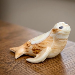 May include: A smooth, polished ceramic figurine of a seal, featuring a cream-colored base with swirling light brown and tan marbled patterns. The seal has dark, almond-shaped eyes and a sleek, rounded body, captured in a resting, upright pose.