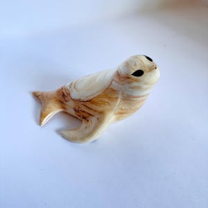 May include: A ceramic figurine of a seal with a glossy finish. The body features a cream-colored base with swirling light brown and tan wood-grain patterns. It has two dark, almond-shaped eyes and a small nose, depicted in a resting position.