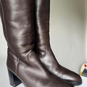 Puede incluir: Un par de botas altas de cuero marrón oscuro con acabado liso, punta redondeada y un tacón de bloque resistente. Las botas presentan costuras visibles en los paneles laterales y una suela gruesa de goma negra texturizada para mayor agarre.
