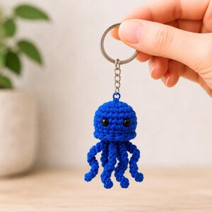 May include: A bright blue crocheted jellyfish keyring featuring a rounded head with two small black bead eyes and several dangling, curled tentacles. The textile figure is attached to a silver-toned metal split ring by a short chain link.