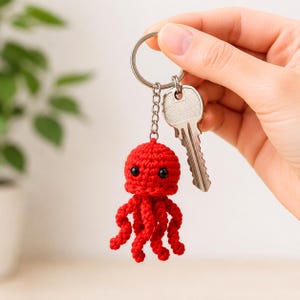 May include: A bright red crocheted octopus keyring featuring a round head with two small black bead eyes and eight dangling tentacles. The octopus is attached to a silver metal split ring and chain, which also holds a single silver house key.
