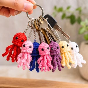May include: A metal keyring holding seven small, crocheted octopus charms in red, light pink, blue, medium pink, purple, yellow, and white, each featuring two black bead eyes and dangling tentacles, attached to a silver ring with several metal keys.