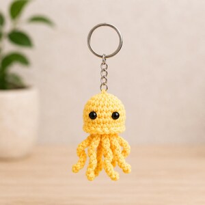 May include: A bright yellow crocheted octopus keyring featuring a rounded head, eight dangling curled tentacles, and two small black bead eyes. The octopus is attached to a silver-coloured metal split ring by a short chain link connector.