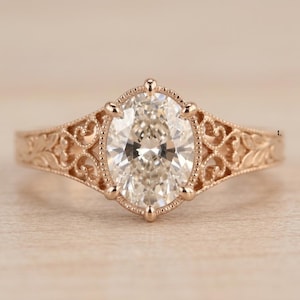May include: A rose gold ring featuring a central oval-cut clear gemstone held by a prong setting. The band is crafted with intricate filigree metalwork and leaf-like engravings extending from the centre stone along the sides of the ring.