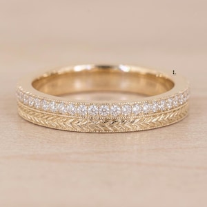 May include: A gold ring featuring a row of small, round, clear stones set along the top edge, complemented by a detailed, engraved leaf-like pattern running horizontally along the lower band, creating a textured, layered appearance.
