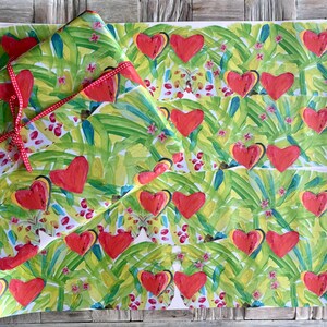 May include: Gift wrap featuring a vibrant pattern of red hearts and green foliage. The paper is tied with a red and white polka dot ribbon. The design includes small pink floral accents, creating a cheerful and festive look.