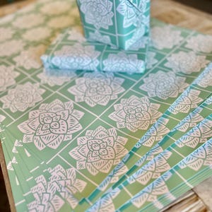 May include: A stack of mint green wrapping paper sheets featuring a repeating white grid pattern with a stylized white succulent or flower illustration in each square. A small box wrapped in the same paper sits on top of the flat sheets.