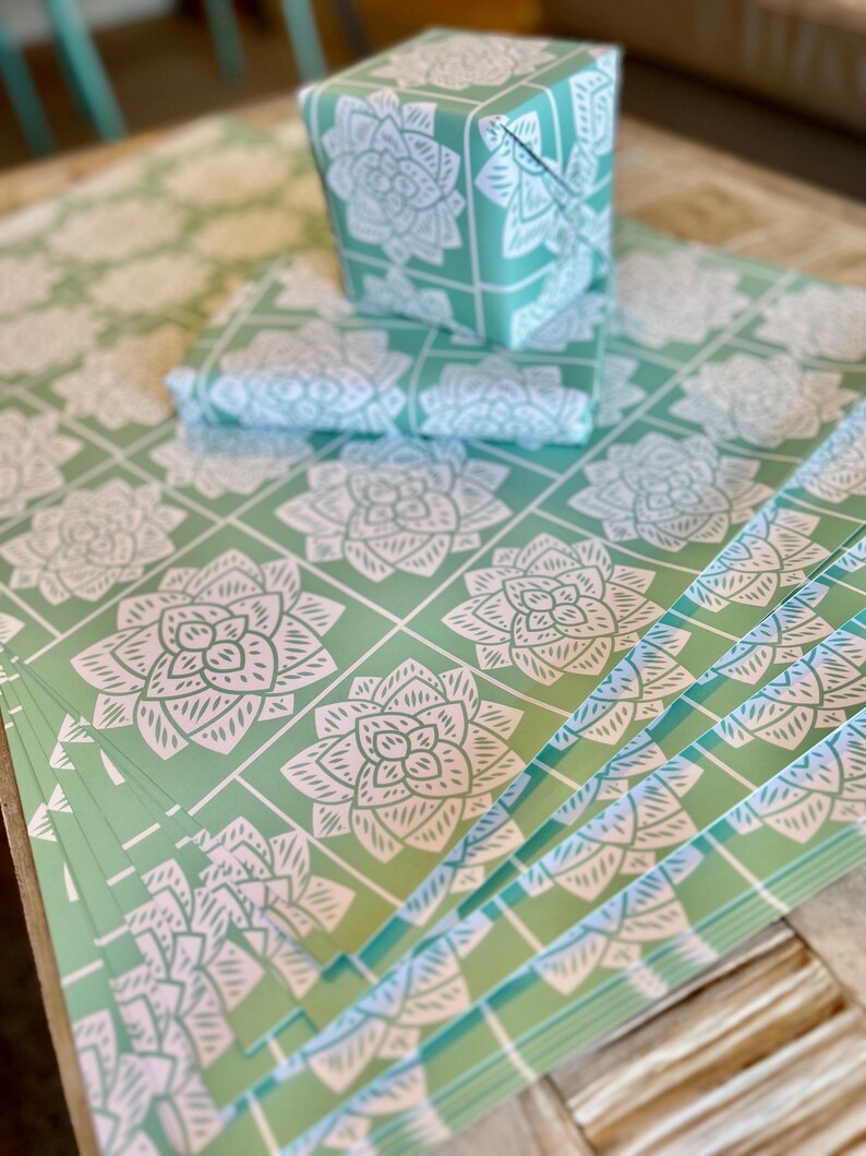 May include: A stack of mint green wrapping paper sheets featuring a repeating white grid pattern with a stylized white succulent or flower illustration in each square. A small box wrapped in the same paper sits on top of the flat sheets.