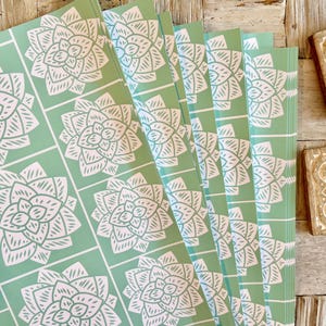 May include: A stack of rectangular sheets of paper featuring a repeating grid pattern of white succulent illustrations on a light mint green background. The paper sheets are arranged in a slightly fanned-out overlapping formation.