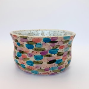 Puede incluir: Un cuenco de cerámica con una base de color crema moteada que presenta un patrón de pinceladas horizontales en tonos azul, rosa, morado y verde oliva. El interior del cuenco también está salpicado de pequeños puntos oscuros.