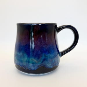 May include: A ceramic mug with a glossy finish featuring a gradient color scheme that transitions from deep black at the top and bottom to vibrant shades of purple, royal blue, and turquoise in the center, complete with a curved handle.
