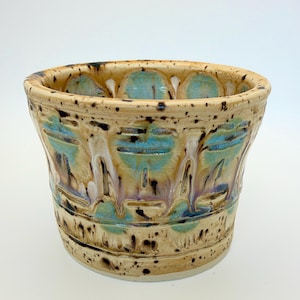 May include: A ceramic planter with a flared rim, featuring a tan base glaze with speckled dark brown accents. The exterior is decorated with vertical carved channels filled with a glossy turquoise and purple drip glaze pattern.