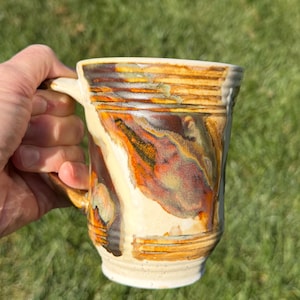 May include: A ceramic mug featuring a variegated glaze with swirling patterns of cream, burnt orange, deep brown, and hints of sage green. The cylindrical vessel has a textured surface with horizontal ridges near the rim and base.