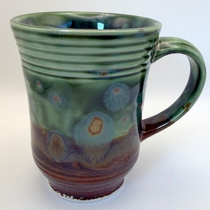 May include: A ceramic mug featuring a ribbed upper section and a smooth lower body. The glaze transitions from a deep reddish-brown at the base to a variegated sage green at the top, accented with crystalline patterns resembling small, pale floral bursts.