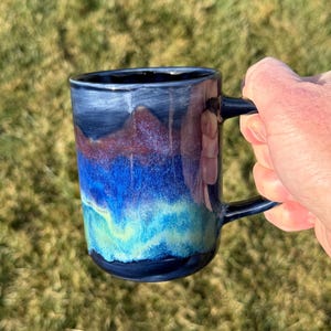 May include: A ceramic mug featuring a multi-colored glaze with horizontal bands of dark navy blue, deep maroon, vibrant royal blue, and bright turquoise. The mug has a smooth, glossy finish and a dark navy blue handle on the right side.