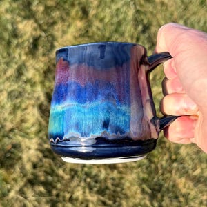 May include: A ceramic mug featuring a glossy glaze with blended horizontal bands of deep navy blue, vibrant royal blue, turquoise, and hints of purple. The mug has a smooth, rounded body and a curved handle on the right side.
