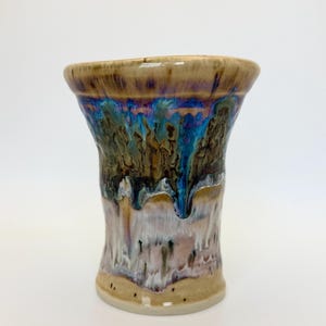 May include: A ceramic tumbler featuring a flared rim and a cylindrical body. The glaze displays a layered effect with a tan base, transitioning into a dark brown and vibrant blue drip pattern that cascades down the upper section of the vessel.