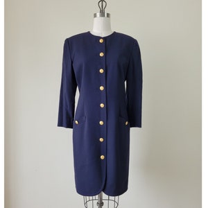 May include: A navy blue dress with long sleeves and a button-up front. The dress features gold-colored buttons and two front pockets. The dress is displayed on a mannequin.