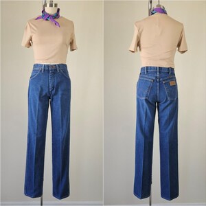 May include: A vintage outfit featuring a tan short-sleeved top and high-waisted, straight-leg blue jeans. A patterned scarf is tied around the mannequin's neck. The jeans have a classic five-pocket design and a brown patch on the back.