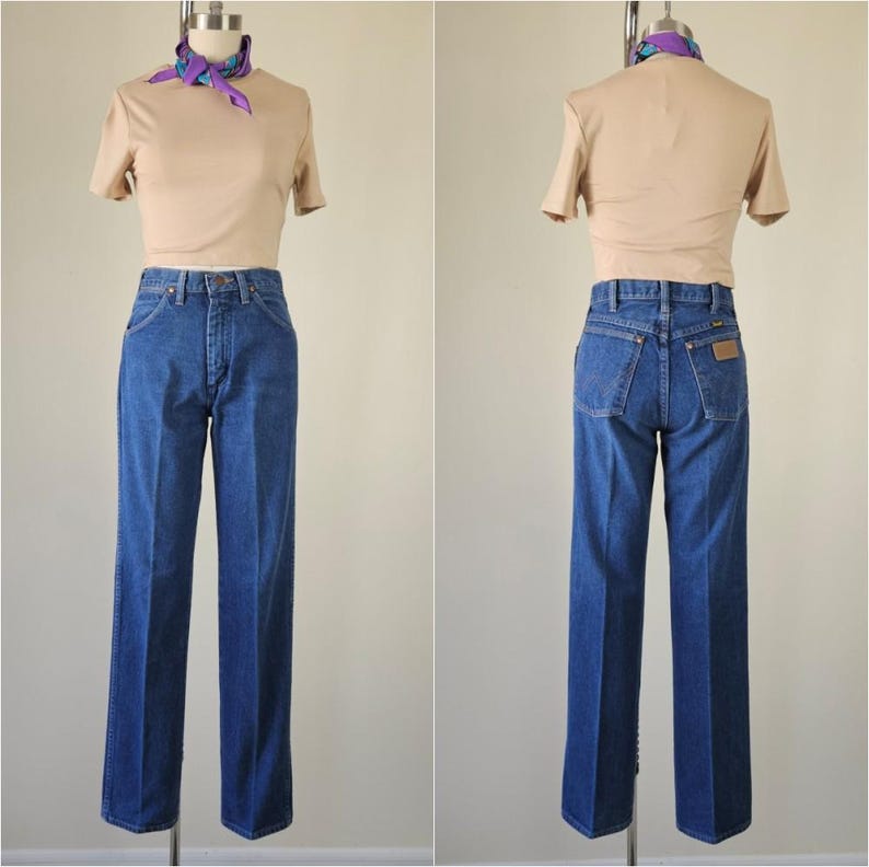 May include: A vintage outfit featuring a tan short-sleeved top and high-waisted, straight-leg blue jeans. A patterned scarf is tied around the mannequin's neck. The jeans have a classic five-pocket design and a brown patch on the back.