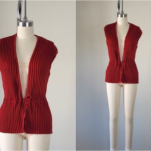 May include: A red knitted sleeveless vest with a tie at the waist.